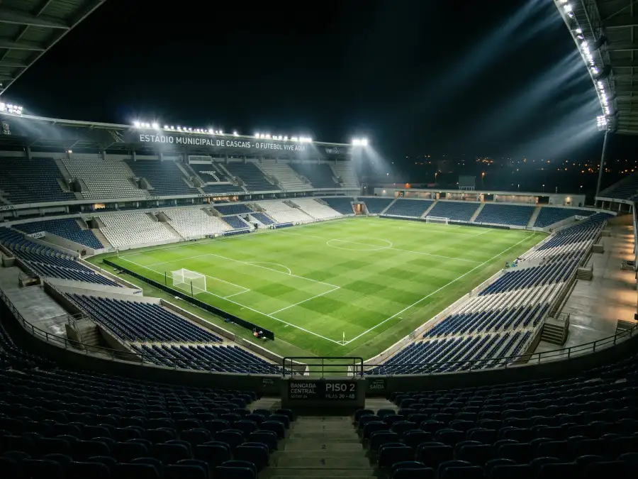Campo de futebol europeu iluminado à noite com relva verde em destaque e bancadas em fundo